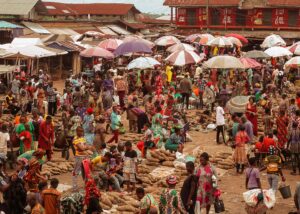 Crowded outdoor market scene with colorful clothing, stalls, and numerous people under striped and solid umbrellas.