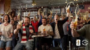 Crowd of excited fans in a bar cheering with raised beer glasses as liquid splashes in the air, American flag on wall behind them.