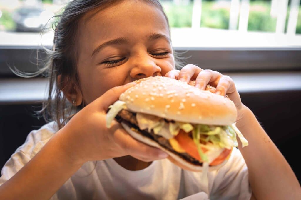 Young girl taking a big bite of a sesame-seed hamburger with lettuce and tomato at a table