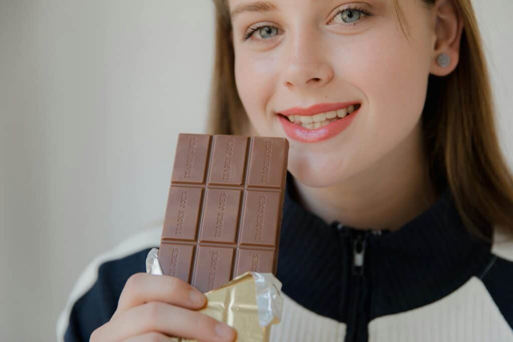 Smiling young woman holding a large milk chocolate bar close to her mouth.