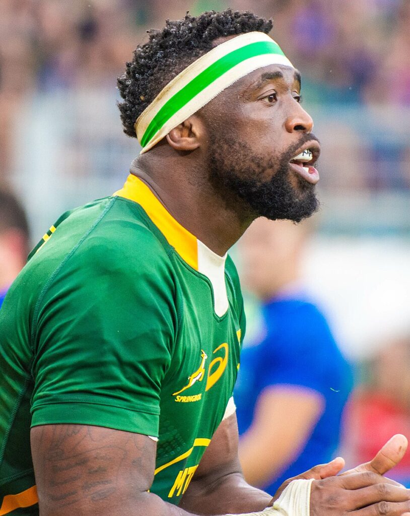 Close-up of a rugby player in a green jersey with a green-white headband, shouting or calling out on the field.