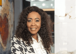 Portrait of a smiling woman with curly hair, wearing a black-and-white geometric blazer and white blouse, in a modern office setting.