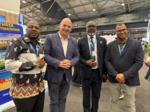 Four men posing at a convention booth; two in suits, one in a colorful patterned shirt, all wearing lanyards, with exhibition booths and ceiling rigging behind them.