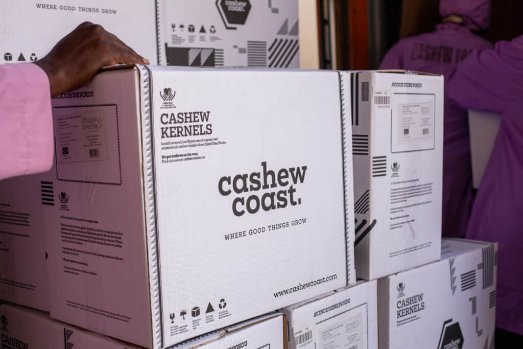 Boxes labeled Cashew Coast being stacked, with a person in a pink sleeve touching the top box, in a warehouse setting.