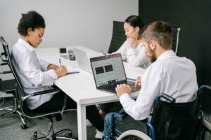 Three coworkers collaborate at a white conference table; two use laptops, one writes notes, and a person in a wheelchair sits at the end of the table.