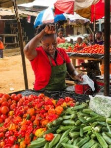 Market vendor in a red shirt and green apron handling peppers and tomatoes at a bustling outdoor stall.