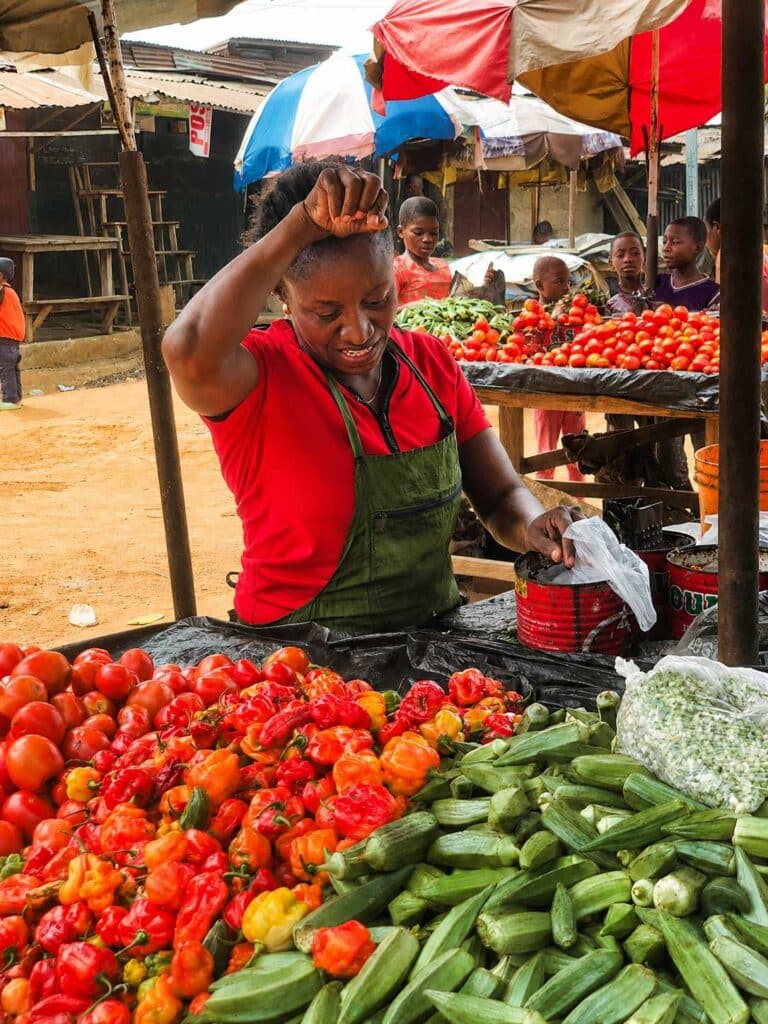 Market vendor in a red shirt and green apron handling peppers and tomatoes at a bustling outdoor stall.