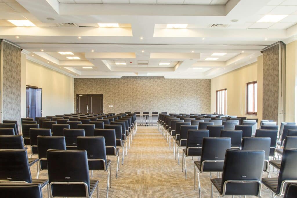 Empty conference hall with many black chairs arranged in rows facing a front stage area.