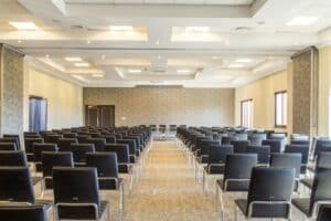 Empty conference hall with many black chairs arranged in rows facing a front stage area.