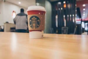 Starbucks hot coffee in a paper cup with sleeve on a wooden table inside a cafe, blurred figure in the background