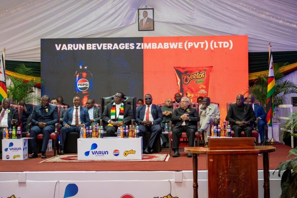 Panel of executives seated on stage at a corporate event; large screen behind reads VARUN BEVERAGES ZIMBABWE (PVT) (LTD) with snack imagery like Pepsi and Cheetos, and flags around the stage.