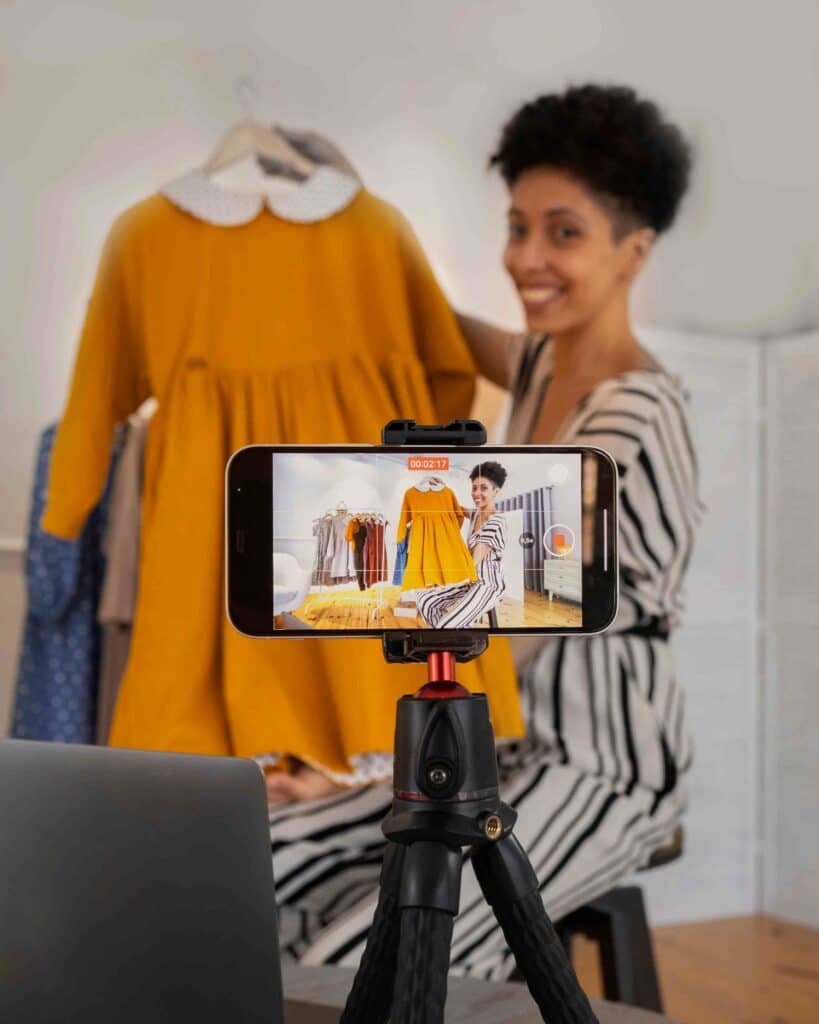 Person films clothing on a tripod-mounted smartphone, with an orange dress in the foreground on a rack in a shop setting piece together a fashion video