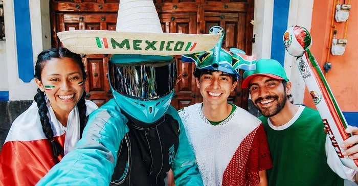 Four friends pose for a selfie in Mexico-themed outfits: teal helmet and gear, sombrero with a 'MEXICO!!' banner, and festive face paint/flags.