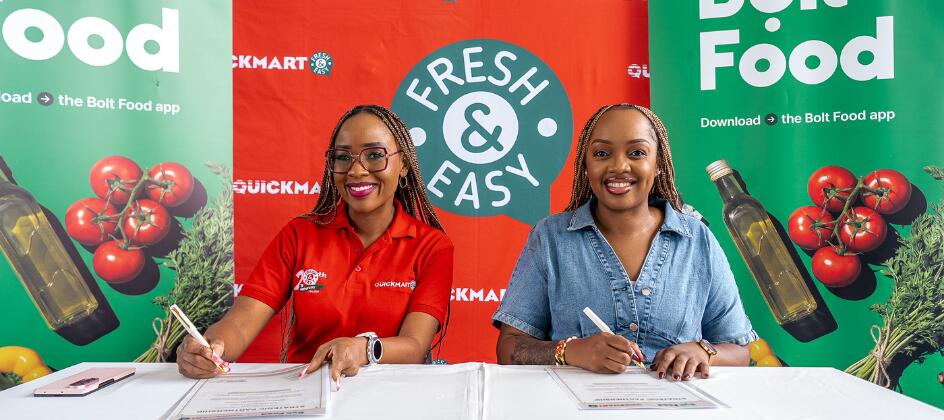Two women sit at a table signing documents at a Bolt Food promotional booth, with a Fresh & Easy backdrop featuring tomatoes and olive oil imagery.