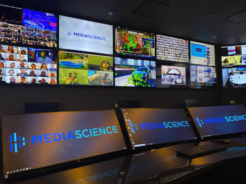 Control room with a wall of video screens displaying a Mediascience logo and live feeds, plus three desk monitors showing the same logo.
