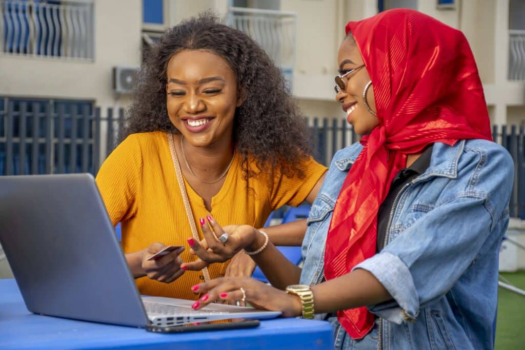 Two women sit at a blue table outdoors, one in a mustard top using a laptop while the other in a red headscarf smiles at her.