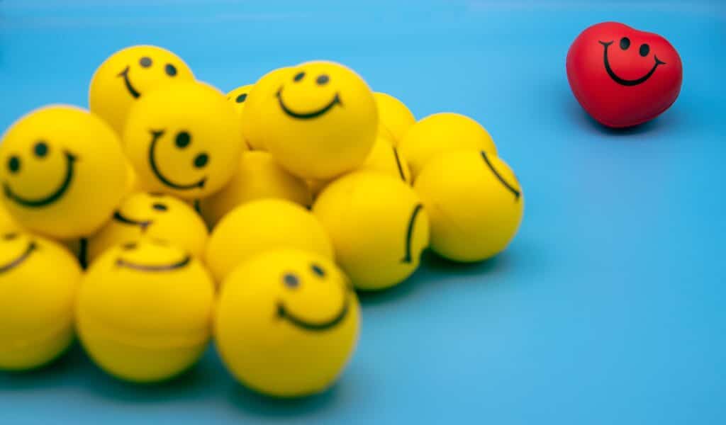 Cluster of yellow smiley-face balls on a blue surface with one red smiling ball away from the group.