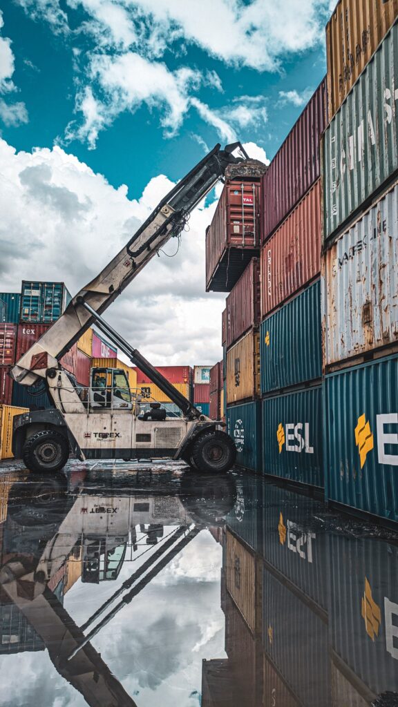 Crane lifting a red shipping container among stacked colorful containers in a port yard under a blue sky with clouds, reflecting on wet pavement.