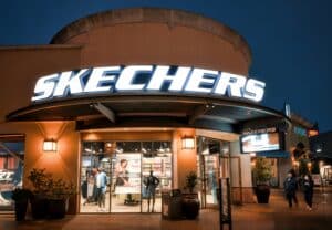 Skechers storefront at night with a bright illuminated sign and glass displays showing mannequins and clothing outside the store in warm lighting.