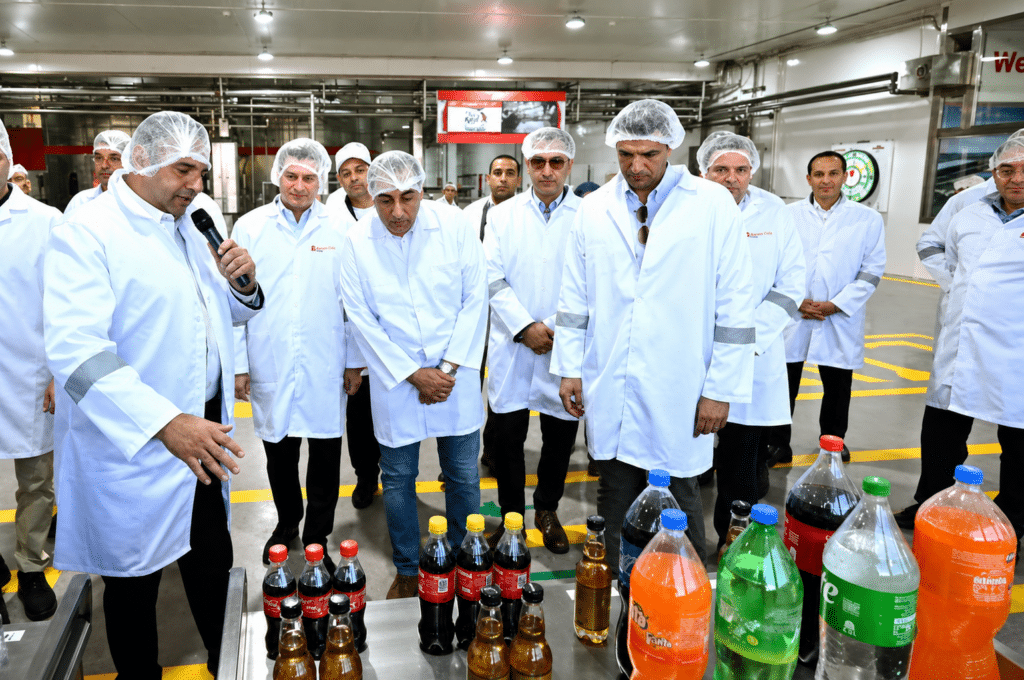 Group of factory workers in lab coats and hairnets listen as a presenter speaks near a conveyor with assorted soda bottles on a production line.