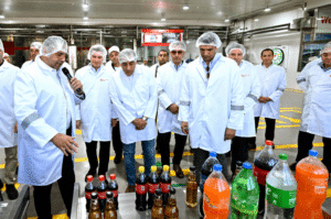 Group of factory workers in lab coats and hairnets listen as a presenter speaks near a conveyor with assorted soda bottles on a production line.