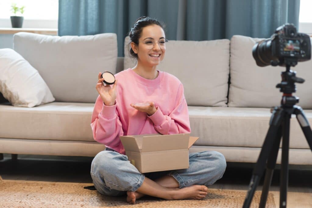 Smiling woman in a pink sweatshirt sits on the floor at home, holding an open makeup compact and a small container while filming with a camera on a tripod nearby.