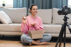Smiling woman in a pink sweatshirt sits on the floor at home, holding an open makeup compact and a small container while filming with a camera on a tripod nearby.