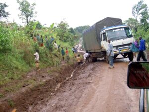 Mud-slick rural road; a large white truck with a blue tarp stuck in the mud while villagers gather nearby.
