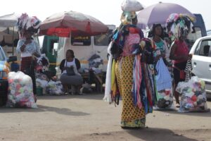 Women in a busy outdoor market balance colorful clothing bundles on their heads beside parked vans and tents.