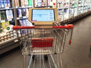 Shopping cart with a red handle and a mounted touchscreen displaying a store map in a supermarket aisle within a grocery store