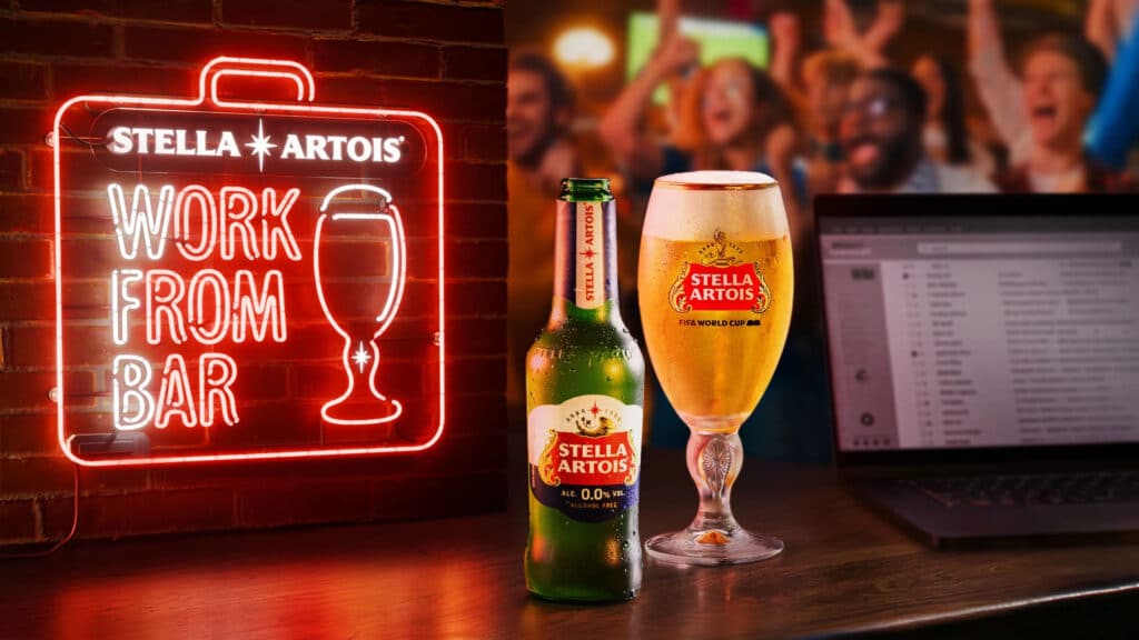 Neon sign says 'Work From Bar' beside a Stella Artois bottle and beer glass on a bar counter with a blurred crowd in the background.