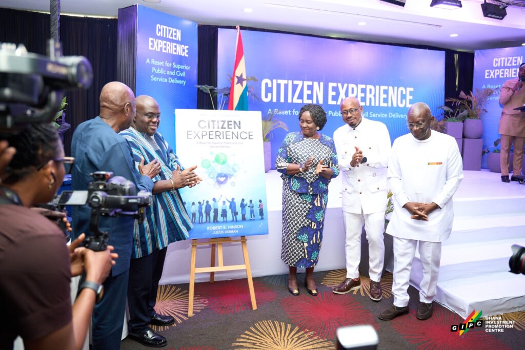 Dignitaries and photographers at a 'Citizen Experience' event on a blue stage backdrop with a poster on an easel nearby.