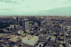 15 July - Urbanisation – African Marketing Confederation – African Marketing Confederation Aerial view of a coastal city with tall skyscrapers, dense low-rise blocks, and a distant shoreline on the horizon.