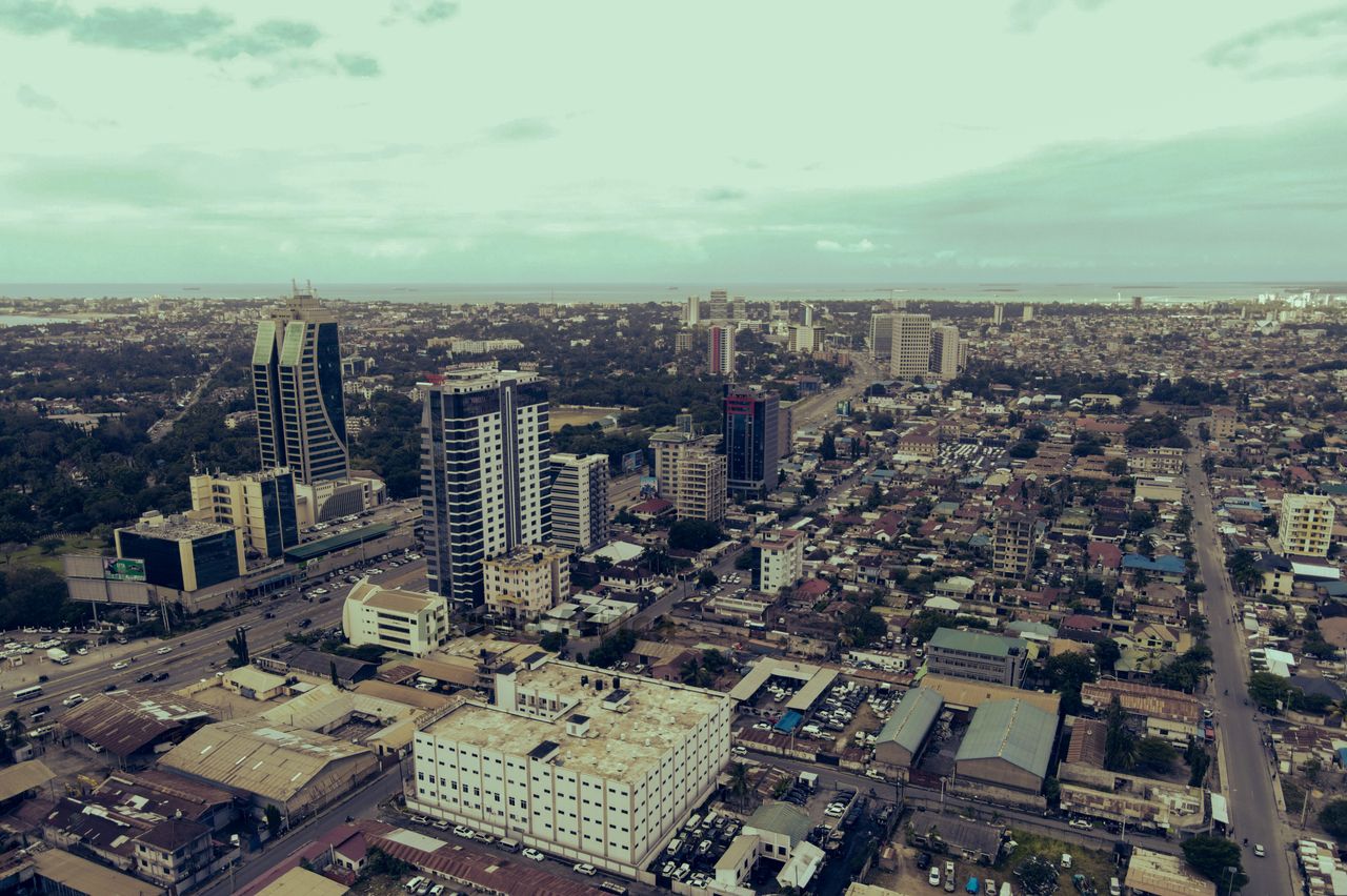 Aerial view of a coastal city with tall skyscrapers, dense low-rise blocks, and a distant shoreline on the horizon.