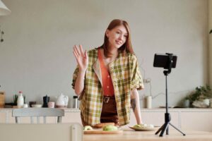 17 July - Social Media – African Marketing Confederation – African Marketing Confederation Woman in a plaid jacket waves at a smartphone on a tripod while filming in a bright kitchen with food on the table behind her