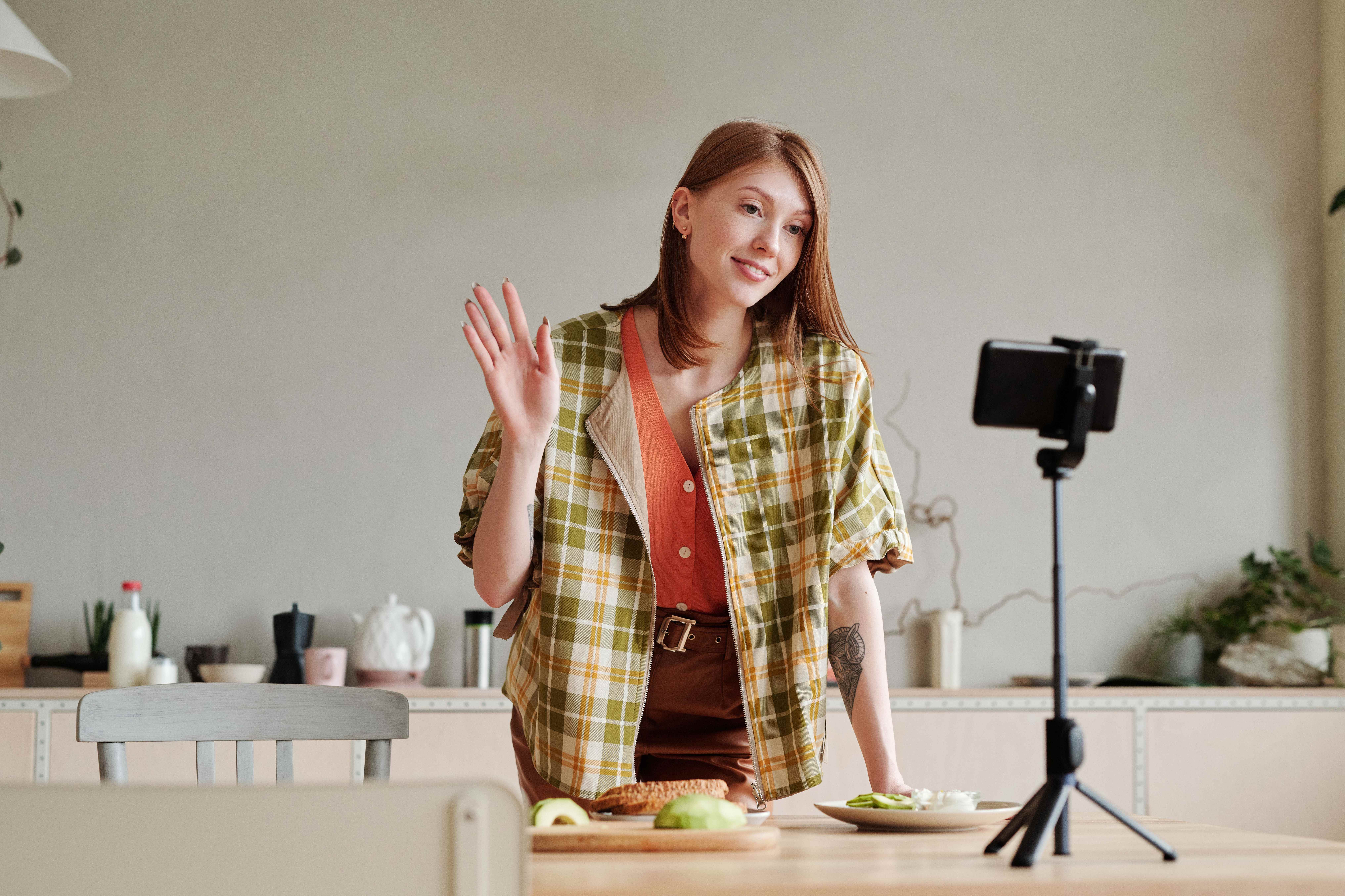 Woman in a plaid jacket waves at a smartphone on a tripod while filming in a bright kitchen with food on the table behind her