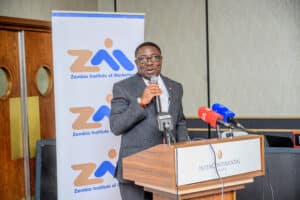 Man in a dark suit speaks into a microphone at a press conference, with a Zambia Institute of Marketing banner behind him and several microphones on the podium desk behind him.