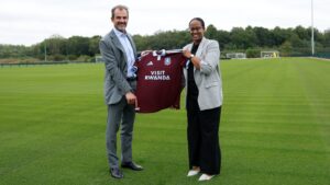 20 July - Sport Sponsorship – African Marketing Confederation – African Marketing Confederation Two adults in business attire stand on a soccer field, holding a maroon jersey that reads 'Visit Rwanda'.
