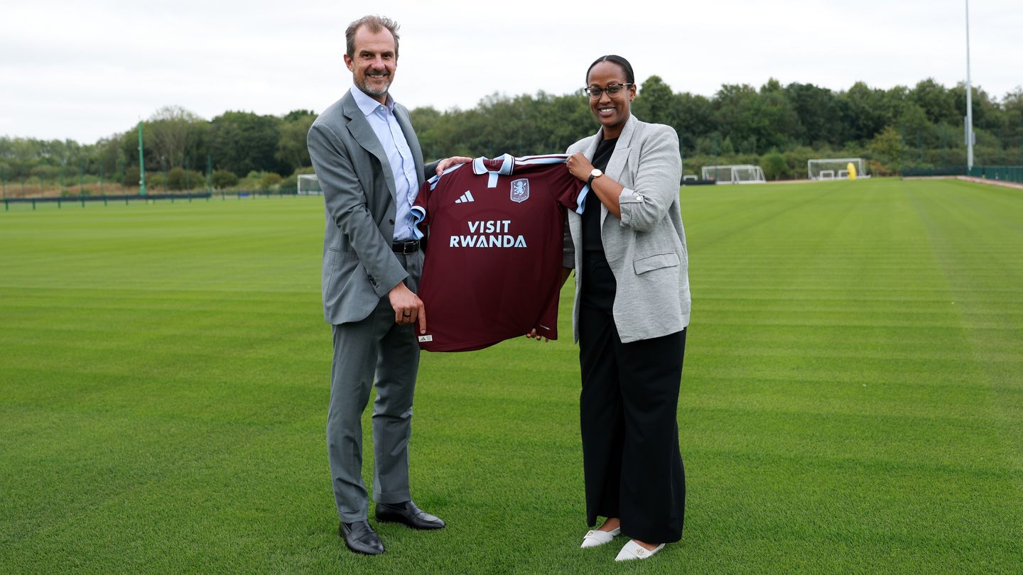 Two adults in business attire stand on a soccer field, holding a maroon jersey that reads 'Visit Rwanda'.