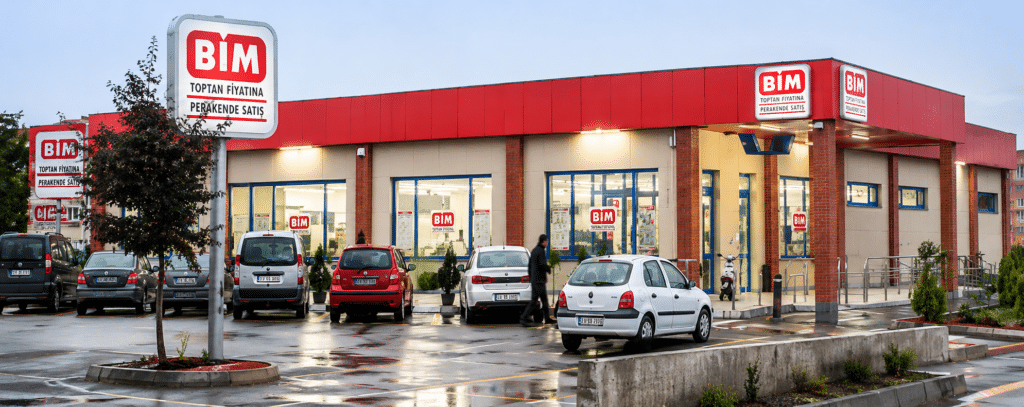 Exterior of a BIM store with red signage and blue-framed windows, parked cars in front on a wet lot.