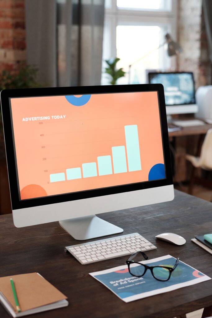 Desk with a computer monitor showing an orange bar chart labeled 'Advertising Today', plus keyboard, mouse, and glasses on a blue magazine.