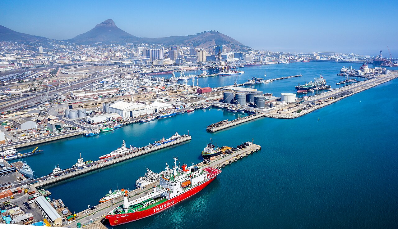 22 July - Supply Chain – African Marketing Confederation – African Marketing Confederation Busy port with cranes, warehouses, and a red training ship docked at the quay; city and hills in the background.
