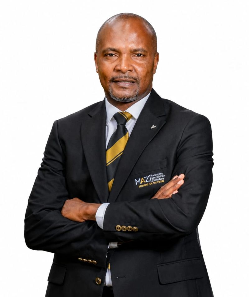 Professional portrait of a middle-aged man in a black suit, white shirt, and gold-and-black striped tie, arms crossed, against a white background; blazer features a small logo.
