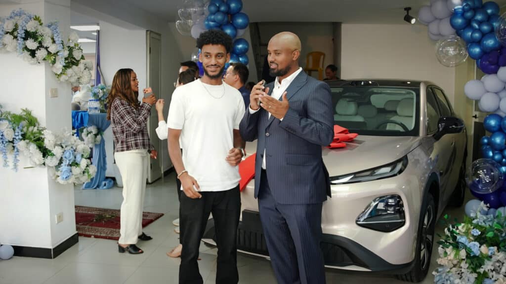 Salesperson in a suit explains features to a man in a white T-shirt beside a new silver car in a showroom with blue balloons and flowers.
