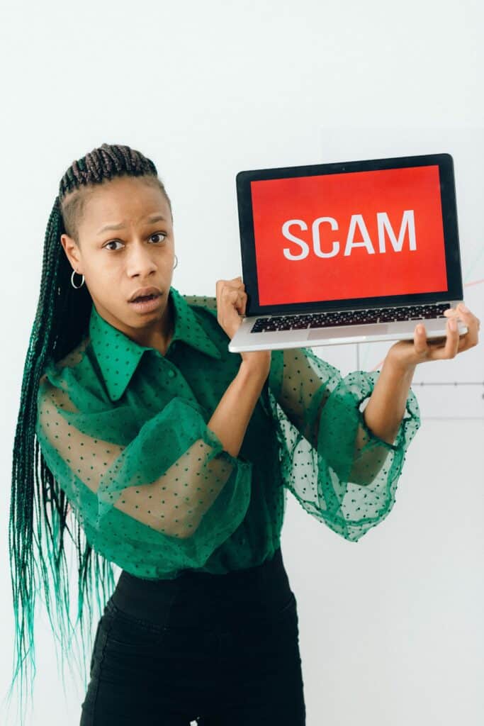 Woman with surprised expression holds laptop showing a red screen reading 'SCAM'.