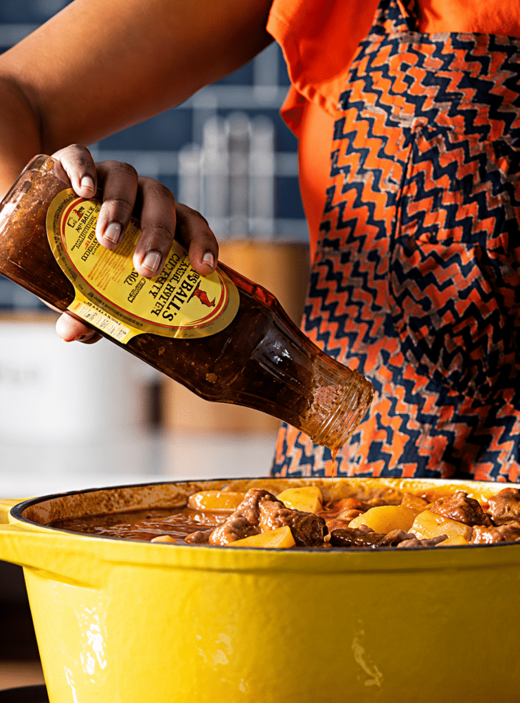Person in an orange patterned apron tilts a bottle of sauce into a large yellow pot of beef stew with potatoes.