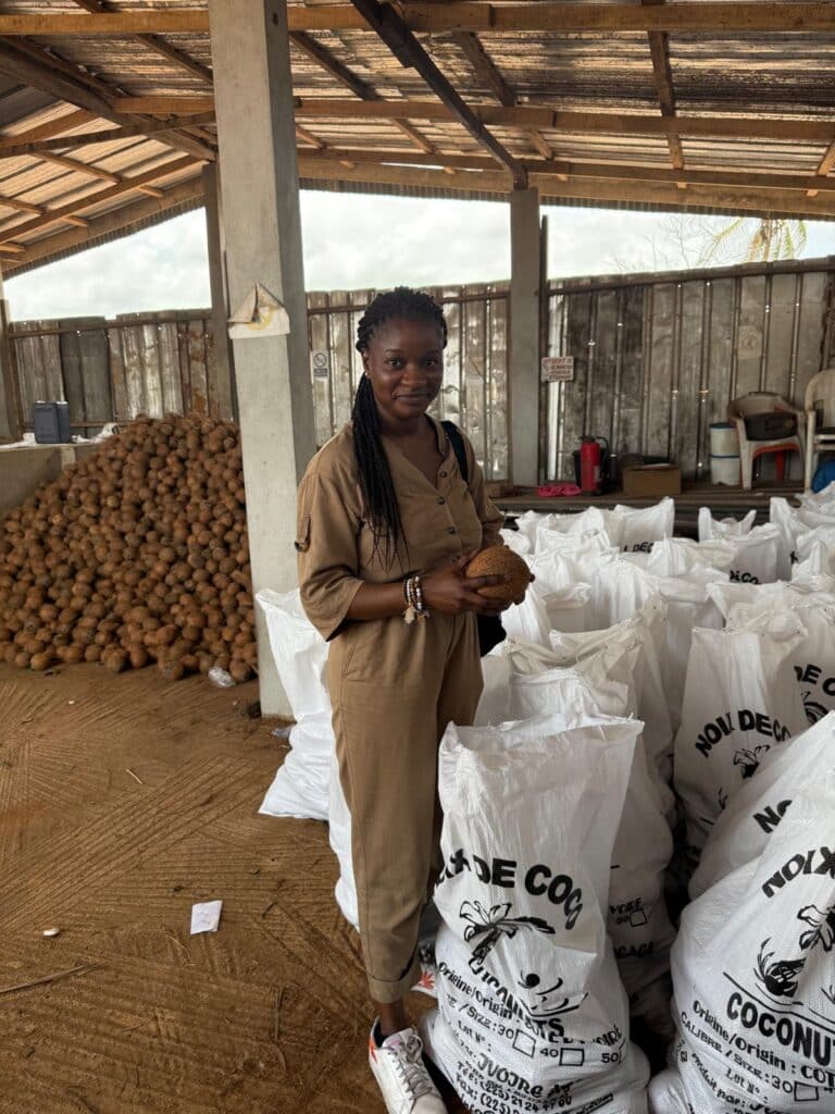 Woman in a tan jumpsuit stands in a warehouse filled with white coconut sacks, holding a coconut and smiling. A large pile of coconuts is behind her under a wooden roof.