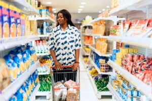 30 July - Shrinkflation – African Marketing Confederation – African Marketing Confederation Woman pushing a shopping cart down a grocery store aisle lined with snack shelves on both sides.