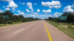 30 July - Supply Chain – African Marketing Confederation – African Marketing Confederation Two green provincial-border signs mark the road crossing between Huila (left) and Cunene (right) provinces under a blue sky with scattered clouds in a rural setting.