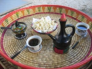 Moroccan-style tea service on a round woven tray: black teapot, patterned lidded pot, two cups of dark tea, and a bowl of popcorn in the center.